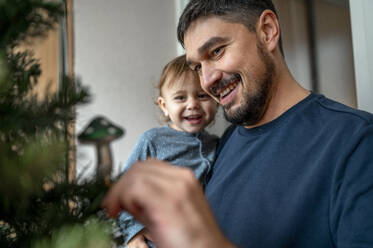 Happy father carrying son and decorating Christmas tree at home - ANAF00330