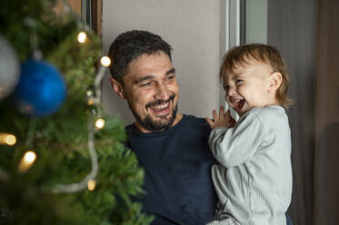 Glücklicher Vater trägt süßen Jungen vor dem Weihnachtsbaum zu Hause - ANAF00339