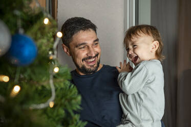 Happy father carrying cute boy in front of Christmas tree at home - ANAF00339