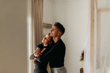 Zärtliches junges Paar, das sich umarmt und an den Händen hält, während es im Haus steht. Verliebtes Paar, das einen romantischen Moment zu Hause erlebt. - JLPSF24896