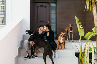 Ein junges Paar küsst sich zärtlich, während es mit seinen Hunden auf der Veranda sitzt. Zwei liebevolle Tierbesitzer verbringen viel Zeit mit ihren liebenswerten Hunden zu Hause. - JLPSF24899