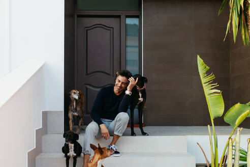 Cheerful pet owner laughing happily while sitting on the porch with all his dogs. Loving man spending some quality time with his family of adorable dogs. - JLPSF24917