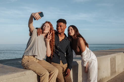 Drei junge Freunde machen ein Gruppen-Selfie am Meer. Eine Gruppe unbeschwerter Freunde posiert für das Fotohandy, während sie am Wochenende zusammen abhängen. Freunde machen gemeinsame Erinnerungen. - JLPSF24923