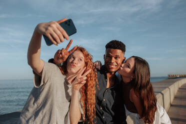 Junge Freunde machen ein Gruppen-Selfie am Meer. Eine Gruppe unbeschwerter Freunde posiert für das Fotohandy, während sie am Wochenende zusammen abhängen. Drei Freunde machen gemeinsame Erinnerungen. - JLPSF24924