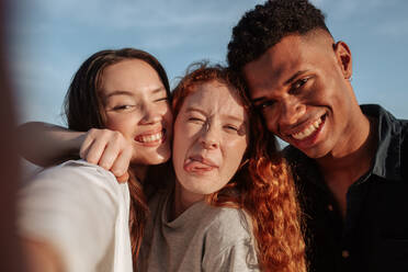 Gen Z-Freunde machen ein Gruppen-Selfie zusammen. Drei multikulturelle Freunde posieren für die Kamera, während sie an einem sonnigen Tag zusammen abhängen. Eine Gruppe junger Menschen, die gemeinsam glückliche Erinnerungen schaffen. - JLPSF24927