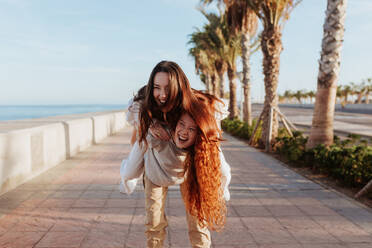 Fröhliche junge Frau, die ihre beste Freundin auf einer Promenade huckepack nimmt. Zwei fröhliche Freundinnen, die zusammen am Wasser Spaß haben. Verspielte Freundinnen, die ihr Wochenende in einer Küstenstadt genießen. - JLPSF24929