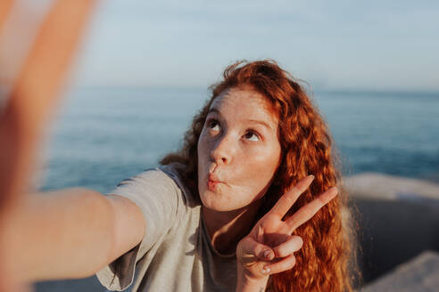 Unbekümmerte junge Frau, die ein Selfie macht, während sie am Meer steht. Junge Frau, die ein lustiges Gesicht macht und das Friedenszeichen zeigt. Fröhliche junge Frau, die Spaß in der Sommersonne hat. - JLPSF24937
