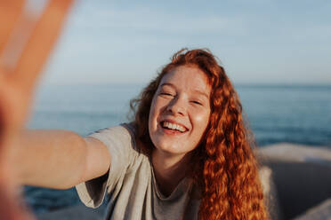 Fröhliche junge Frau, die ein Selfie macht, während sie am Meer steht. Unbekümmerte junge Frau, die glücklich in die Kamera lächelt. Junge Frau im Freien, die Spaß in der Sommersonne hat. - JLPSF24938