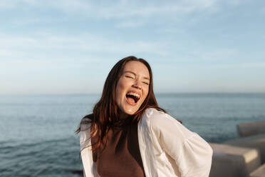 Brünette im Freien, die mit geschlossenen Augen lacht, während sie am Meer steht. Glückliche junge Frau, die sich im Sommer amüsiert. Fröhliche junge Frau, die ein tolles Wochenende am Meer genießt. - JLPSF24942