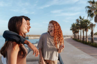 Lachen an der Strandpromenade. Fröhliche junge Frau, die mit ihren beiden Freunden im Freien am Meer lacht. Gruppe von drei multikulturellen Freunden, die am Wochenende eine gute Zeit miteinander verbringen. - JLPSF24946