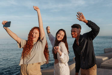 Taking happy selfies by the sea. Group of three friends cheering and dancing in front of a camera phone. Multicultural friends having a good time while hanging out together next to the sea. - JLPSF24951