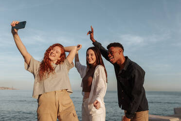 Fröhliche Momente mit Freunden festhalten. Eine Gruppe von drei Freunden, die vor einem Fotohandy jubeln und tanzen. Multikulturelle Freunde haben eine gute Zeit, während sie zusammen am Meer abhängen. - JLPSF24952