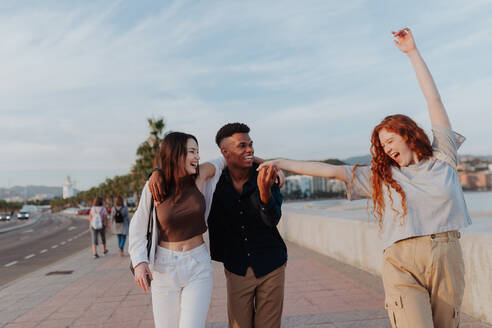 Lebhafte junge Frau tanzt beim Spaziergang mit ihren Freunden. Gruppe von drei glücklichen Freunden, die einen Spaziergang entlang der Promenade machen. Multikulturelle Freunde, die einen Wochenendausflug in einer Küstenstadt genießen. - JLPSF24957