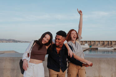 Friends having a good time together outdoors. Group of three happy friends laughing and cheering while standing next to the sea. Multiethnic friends enjoying a weekend getaway in a coastal town. - JLPSF24964