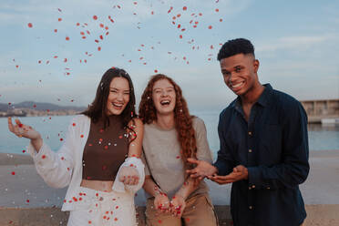 Cheerful friends celebrating red and white confetti outdoors. Group of multicultural friends throwing a confetti party next to the sea. Diverse friends having a good time together on the weekend. - JLPSF24968