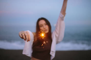 Lassen wir die Dinge funkeln. Unbeschwerte junge Frau, die am Abend am Strand steht und ein funkelndes Licht in der Hand hält. Lebenslustige junge Frau, die mit bengalischen Lichtern feiert. - JLPSF24973