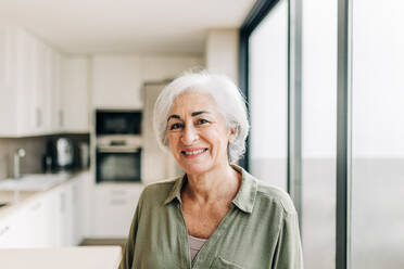 Happy elderly woman smiling at the camera while standing indoors. Cheerful senior woman enjoying her retirement years inside her home. - JLPSF24977