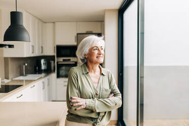 Cheerful senior woman looking away thoughtfully while standing in her home. Happy elderly woman smiling while reflecting on memories of the past. - JLPSF24978