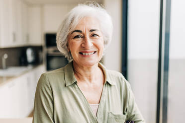 Ältere Frau lächelt in die Kamera, während sie in ihrem Haus steht. Fröhliche ältere Frau, die ihren Ruhestand in ihrem Haus genießt. - JLPSF24980