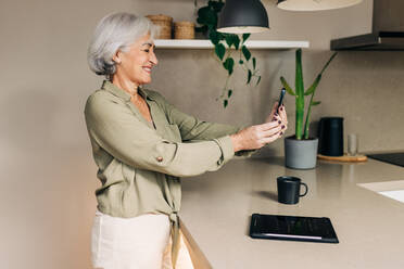 Grauhaarige Seniorin lächelt fröhlich, während sie zu Hause einen Videoanruf entgegennimmt. Fröhliche Seniorin, die mit ihren Lieben über drahtlose Technologie kommuniziert. - JLPSF24989