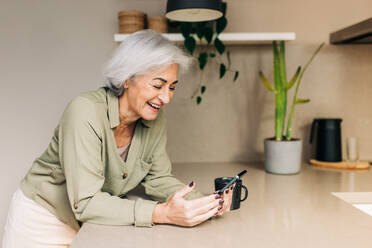 Cheerful senior woman laughing happily while speaking on a video call at home. Grey-haired senior woman communicating with her loved ones using wireless technology. - JLPSF24990