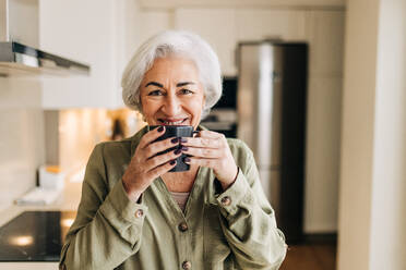 Glückliche ältere Frau, die ihren Tag mit einer Tasse Kaffee beginnt. Fröhliche ältere Frau, die in die Kamera lächelt, während sie in ihrer Küche zu Hause steht. - JLPSF24993