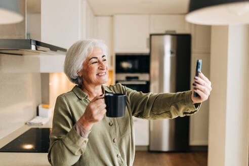 Grauhaarige Seniorin, die zu Hause einen Videogespräch auf ihrem Smartphone führt. Ältere Frau im Ruhestand, die fröhlich lächelt, während sie am Wochenende mit ihren Lieben kommuniziert. - JLPSF24997