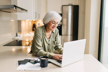 Selbstständige ältere Frau, die zu Hause an ihrem Laptop arbeitet. Glückliche ältere Geschäftsfrau, die eine geschäftliche E-Mail tippt, während sie von zu Hause aus arbeitet. - JLPSF24998