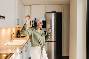 Cheerful senior woman dancing to her favourite music while wearing wireless headphones. Happy elderly woman having a good time while playing music on her smartphone. - JLPSF25001