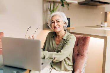 Silver-haired senior businesswoman using a laptop while sitting at her desk. Successful businesswoman smiling happily and enjoying working from home. - JLPSF25005