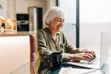 Cheerful senior businesswoman typing on her laptop while sitting at her desk. Successful businesswoman smiling happily and enjoying working from home. - JLPSF25006