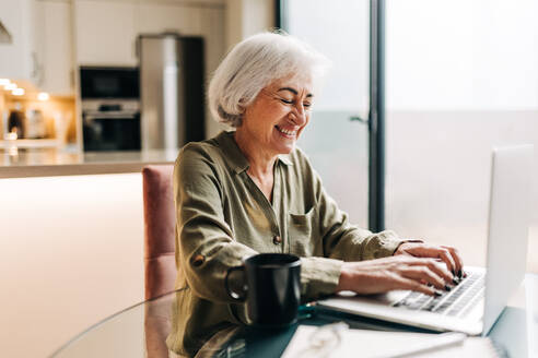 Fröhliche ältere Geschäftsfrau, die an ihrem Schreibtisch sitzend auf ihrem Laptop tippt. Erfolgreiche Geschäftsfrau, die fröhlich lächelt und die Arbeit von zu Hause aus genießt. - JLPSF25006