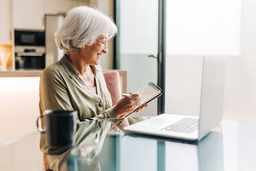 Ältere Geschäftsfrau macht sich während einer Videokonferenz mit ihren Geschäftspartnern Notizen. Ältere Geschäftsfrau lächelt glücklich, während sie an einer Sitzung in ihrem Heimbüro teilnimmt. - JLPSF25007