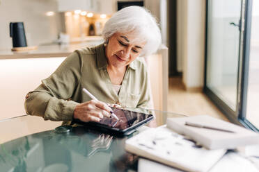 Kreative ältere Frau, die einen Entwurf auf einem digitalen Tablet zeichnet. Ältere Geschäftsfrau, die in ihrem Büro zu Hause an einem neuen Grafikdesign-Projekt arbeitet. - JLPSF25008