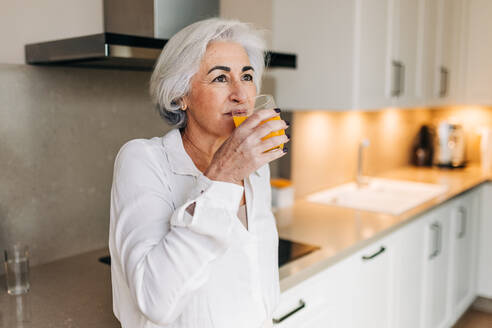 Gesunde ältere Frau, die zu Hause in ihrer Küche ein Glas Fruchtsaft trinkt. Grauhaarige Seniorin, die im Ruhestand auf eine nahrhafte Ernährung achtet. - JLPSF25024