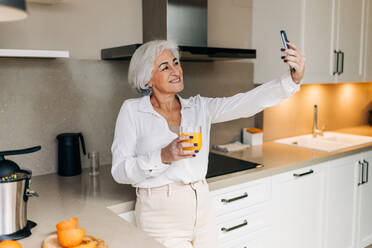 Fröhliche ältere Frau, die einen Videogespräch in ihrer Küche zu Hause führt. Glückliche ältere Frau, die mit ihren Lieben per Video chattet, während sie ein Glas frischen Fruchtsaft genießt. - JLPSF25025