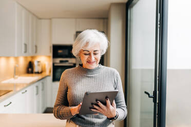Lächelnde ältere Frau, die ein digitales Tablet benutzt, während sie zu Hause steht. Glückliche ältere Frau, die am Wochenende im Internet surft. - JLPSF25027