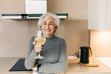 Happy senior woman smiling cheerfully while holding a glass of sparkling wine. Carefree elderly woman celebrating her retirement at home. - JLPSF25032