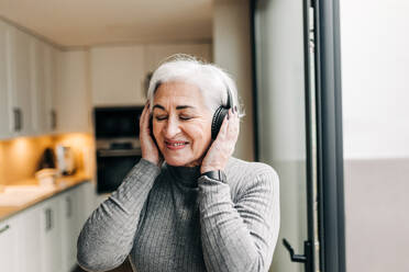 Cheerful elderly woman enjoying listening to her favourite music on wireless headphones. Happy senior woman having fun and enjoying her retirement years at home. - JLPSF25040