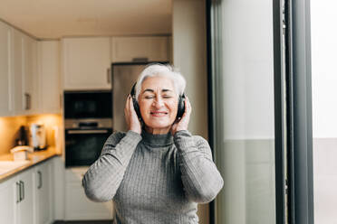 Unbeschwerte ältere Frau, die ihre Lieblingsmusik über drahtlose Kopfhörer hört. Glückliche ältere Frau, die Spaß hat und ihren Ruhestand zu Hause genießt. - JLPSF25041