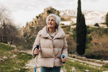 Glückliche ältere Frau beim Trekking mit Wanderstöcken im Freien. Abenteuerlustige ältere Frau lächelt, während sie einen hügeligen Weg hinaufgeht. Reife Frau genießt Freizeitaktivitäten nach der Pensionierung. - JLPSF25048