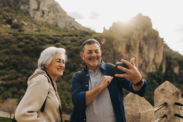Elderly couple having a video call while standing on a hilltop. Happy senior couple enjoying a recreational hike outdoors. Adventurous couple having a good time together after retirement. - JLPSF25059