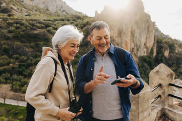 Carefree senior couple having a video call while standing on a hilltop. Happy elderly couple enjoying a recreational hike outdoors. Adventurous couple having a good time together after retirement. - JLPSF25060