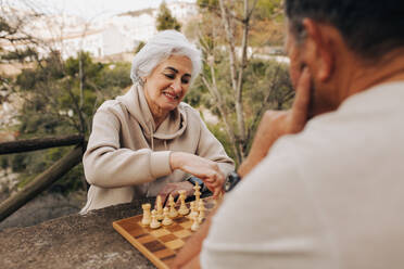 Ein nettes älteres Paar spielt eine Partie Schach in einem Park. Ein glückliches älteres Paar, das nach seiner Pensionierung einige Zeit miteinander verbringt. Ein reifes Paar, das sich im Freien vergnügt. - JLPSF25064