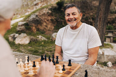 Happy senior man playing chess with his wife in a park. Cheerful elderly couple spending quality time together after retirement. Mature couple enjoying themselves outdoors. - JLPSF25067