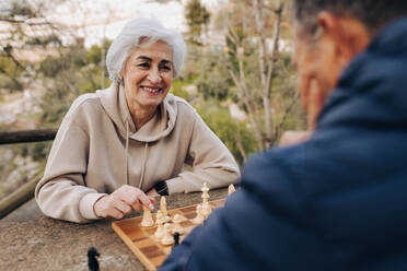 Unbekümmertes älteres Paar, das in einem Park Schach spielt. Glückliches älteres Paar, das nach der Pensionierung einige schöne Stunden miteinander verbringt. Reifes Paar, das sich im Freien amüsiert. - JLPSF25068