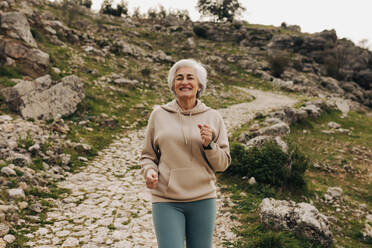 Cheerful senior woman smiling while jogging alone outdoors. Happy elderly woman working out on a hilly trail. Mature woman maintaining a healthy lifestyle after retirement. - JLPSF25073