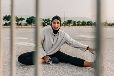 Sportliche Frau mit Hijab, die sich morgens im Freien mit Dehnübungen aufwärmt. Sportliche Muslimin, die in die Kamera schaut, während sie in der Stadt trainiert. - JLPSF25084