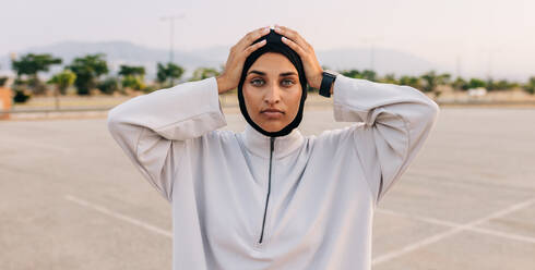 Muslimische Sportlerin, die in die Kamera schaut, während sie mit den Händen auf dem Kopf im Freien steht. Sportliche Frau mit Hijab beim morgendlichen Training. - JLPSF25091