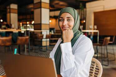 Beautiful Muslim woman looking at the camera while sitting with a laptop in a cafe. Self-employed woman with a hijab doing online freelance work in a restaurant. - JLPSF25101